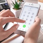 Person holding an iPhone and dialing a number on the phone keypad at a desk with a calculator and plant in the background