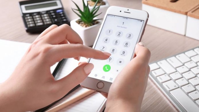 Person holding an iPhone and dialing a number on the phone keypad at a desk with a calculator and plant in the background