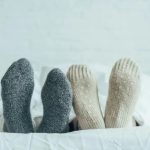 Person lying in bed with gray and beige knitted socks peeking out from under a white duvet.