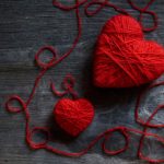 Two red yarn hearts on a dark wooden surface, with loose strands looping around them (Valentine's theme).