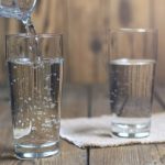 Water is being poured into a tall glass on a wooden table, with a second filled glass nearby and a small towel underneath.