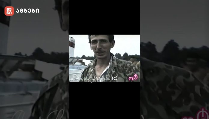 Man in camouflage jacket speaks to the camera outdoors, with watermarked text across the bottom.