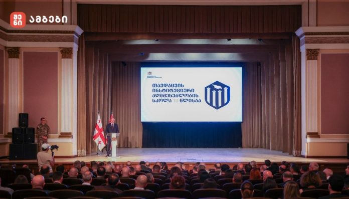 Speaker at a podium on a stage in an auditorium, two Georgian flags beside him and a large screen with a blue emblem and Georgian text behind him