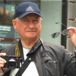 Older man wearing a blue Adidas cap holds a camera in front of him and lifts sunglasses in his right hand.