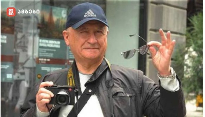 Older man wearing a blue Adidas cap holds a camera in front of him and lifts sunglasses in his right hand.