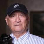 Portrait of an older man in a striped shirt wearing a cap, holding a camera with a Lumix lens.