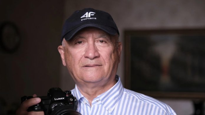Portrait of an older man in a striped shirt wearing a cap, holding a camera with a Lumix lens.