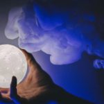 Hand cradling a glowing full-moon orb against a dark blue smoky background.