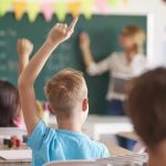 Classroom scene with a teacher at the chalkboard as students raise their hands to answer a question, colorful bunting overhead.