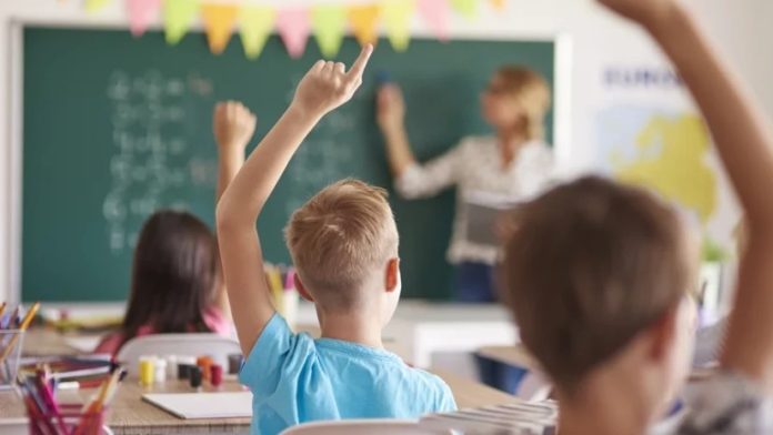 Classroom scene with a teacher at the chalkboard as students raise their hands to answer a question, colorful bunting overhead.