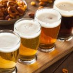 Four beers in a flight lined up on a wooden board with pretzels and nuts nearby, ready for tasting.