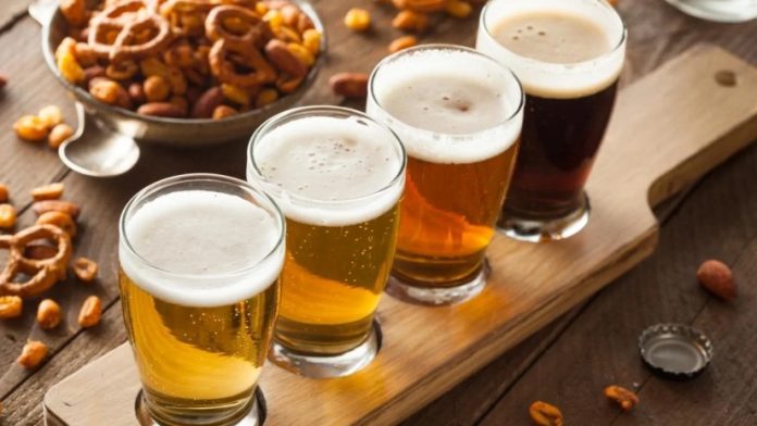Four beers in a flight lined up on a wooden board with pretzels and nuts nearby, ready for tasting.