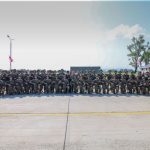 Large group of soldiers in camouflage uniforms posing in a line on a tarmac, with trees and a building in the background.