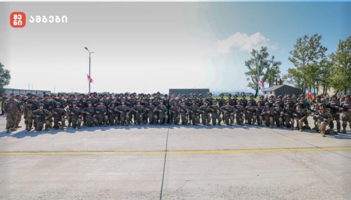 Large group of soldiers in camouflage uniforms posing in a line on a tarmac, with trees and a building in the background.