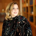 Portrait of a smiling woman with blonde hair wearing a patterned black coat, standing in a library with bookshelves in the background.