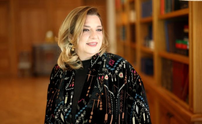 Portrait of a smiling woman with blonde hair wearing a patterned black coat, standing in a library with bookshelves in the background.