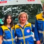 Three emergency responders in blue and yellow uniforms stand in front of a white ambulance with blue markings.