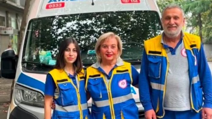 Three emergency responders in blue and yellow uniforms stand in front of a white ambulance with blue markings.