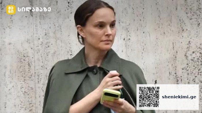 Woman with brown hair in an olive-green coat holding a lime-colored smartphone while standing against a textured concrete wall.