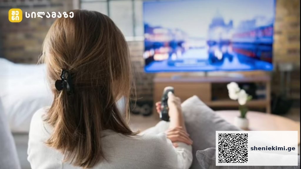 Back view of a woman sitting on a sofa, holding a remote and watching a blurred TV screen in a cozy living room.