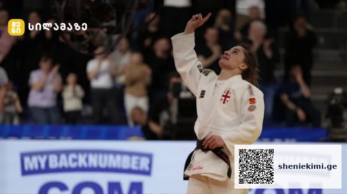 Female judo athlete in a white gi raises her right fist in celebration on the mat as the crowd applauds in the background.