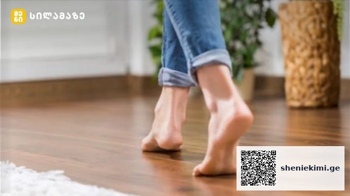 Barefoot person walking on a wooden floor indoors, jeans cuffs rolled up.