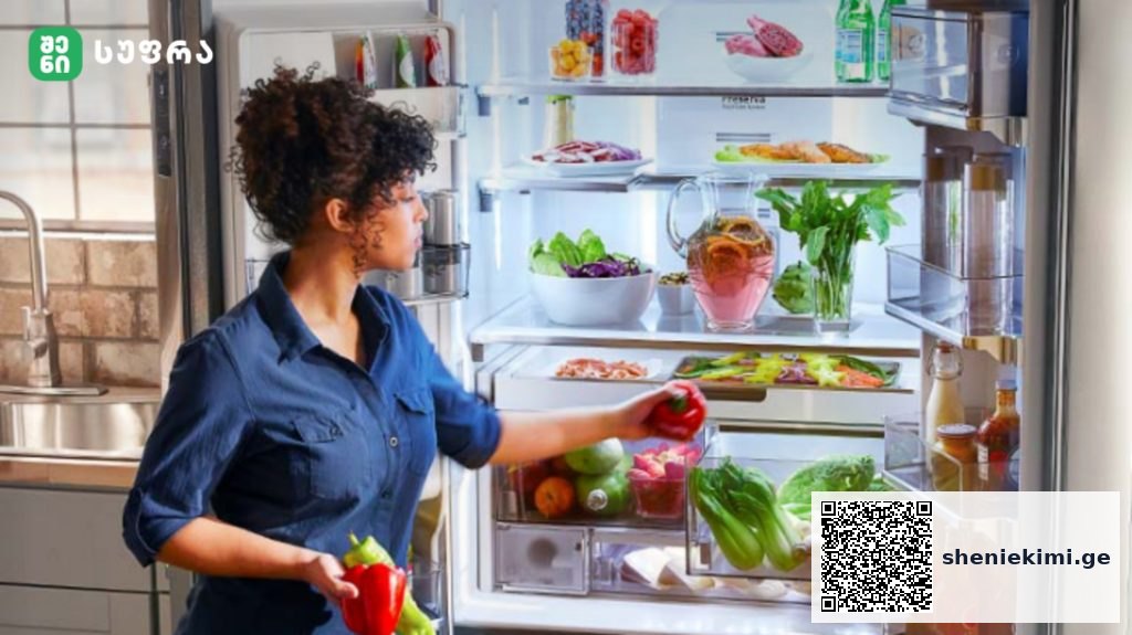 Woman in a blue shirt stands by an open refrigerator, reaching for a red pepper among fresh vegetables and drinks inside the fridge.