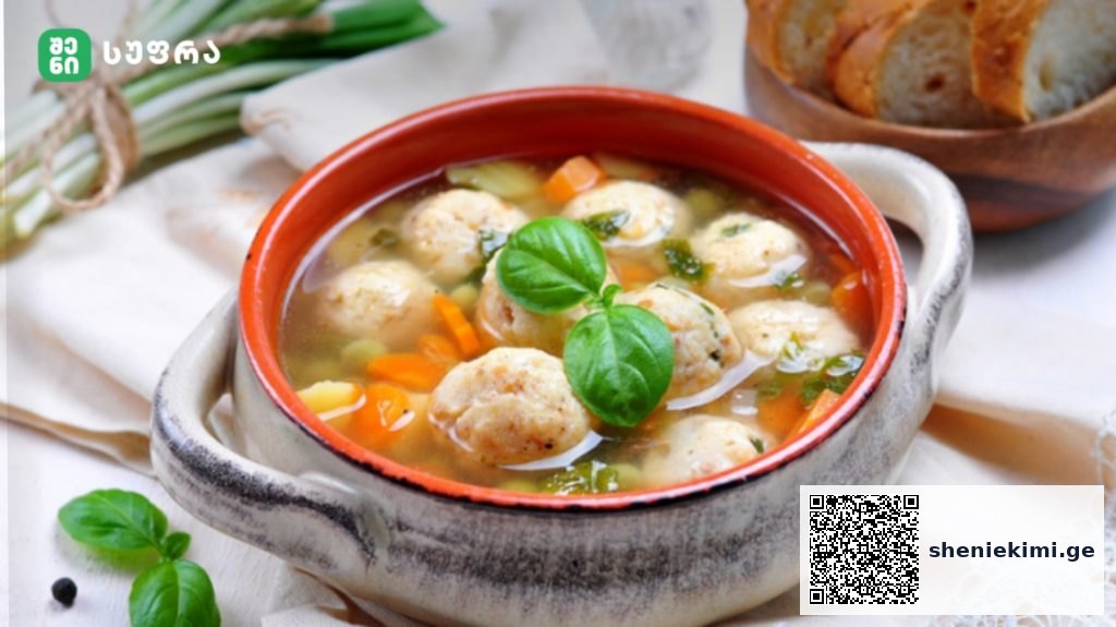 Bowl of meatball soup with carrots and fresh basil, served with bread on a light tablecloth, in a rustic pot.