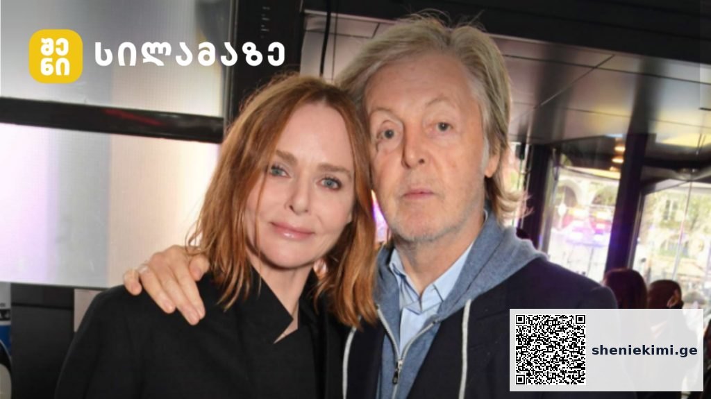 Two adults posing indoors, a woman with red hair and a man with light hair, both looking at the camera; yellow logo in the top-left corner.