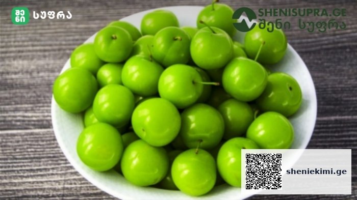 White bowl piled with bright green plums on a dark wooden surface.