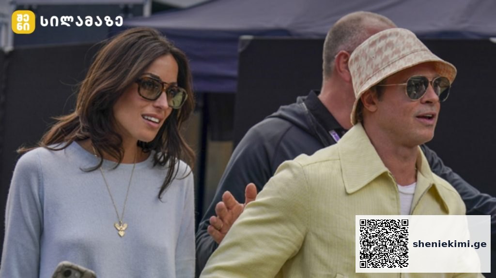 Two stylish adults wearing sunglasses walk outdoors at an event; woman in pale blue top with a heart necklace, man in a pale yellow jacket and bucket hat.