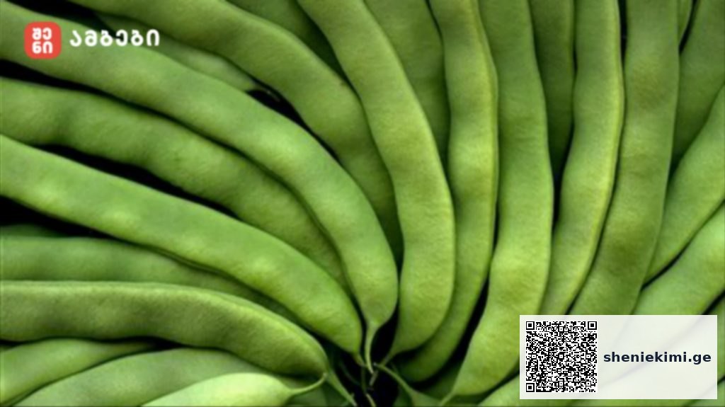 Close-up of fresh green beans stacked in a dense arrangement, with a small red logo in the top-left corner.