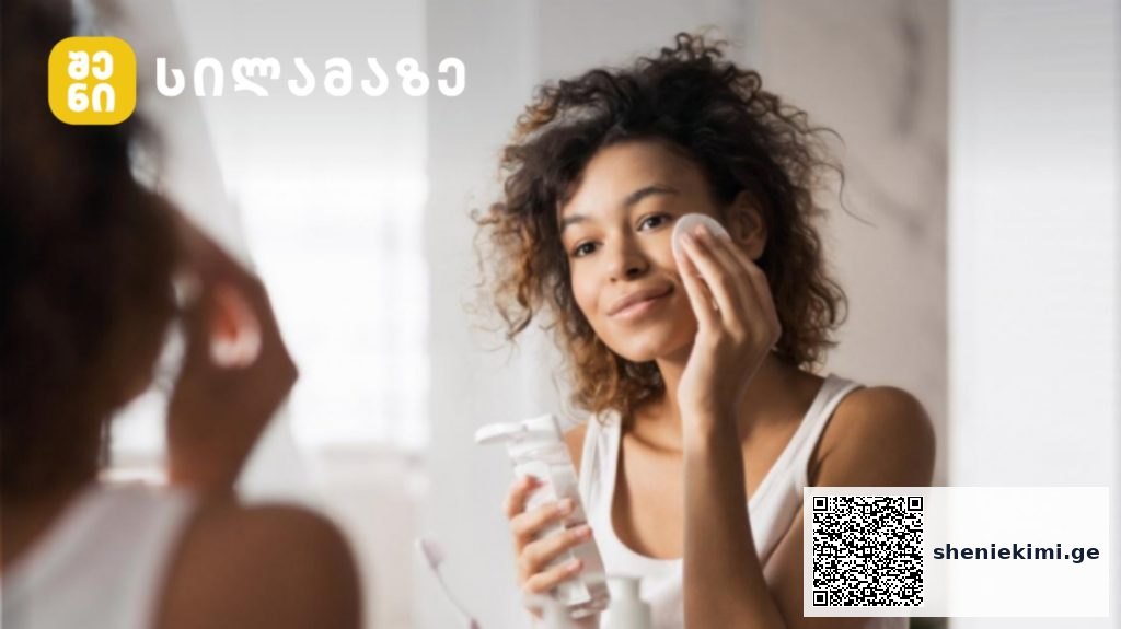 Woman applying skincare to her face while looking in a mirror, holding a cleanser bottle and cotton pad, with a logo in the top-left corner.