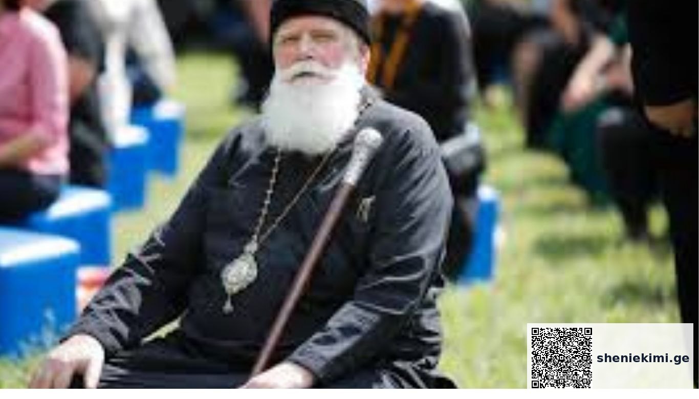 Older man with a long white beard in black robes and a hat, seated outdoors with a staff and a pendant, at a park event. Religious elder in black attire and a medallion, holding a staff at an outdoor gathering with blurred onlookers behind. Senior clergy figure with a white beard, sitting with a walking staff during an outdoor ceremony.