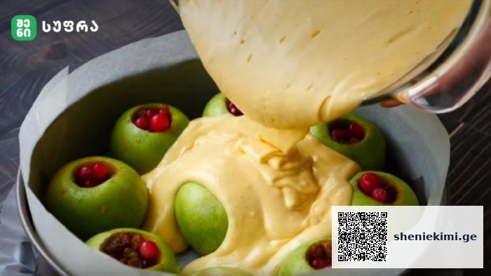 Green apples stuffed with red berries in a round cake pan, as creamy batter is poured over them.
