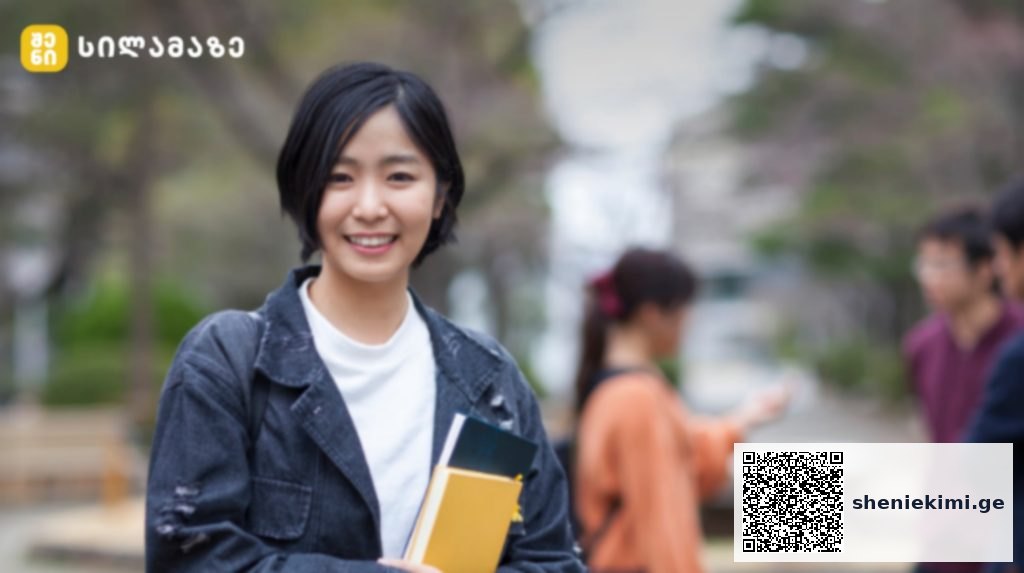Smiling young woman with short black hair holding notebooks on a campus walkway, jacketed in denim.