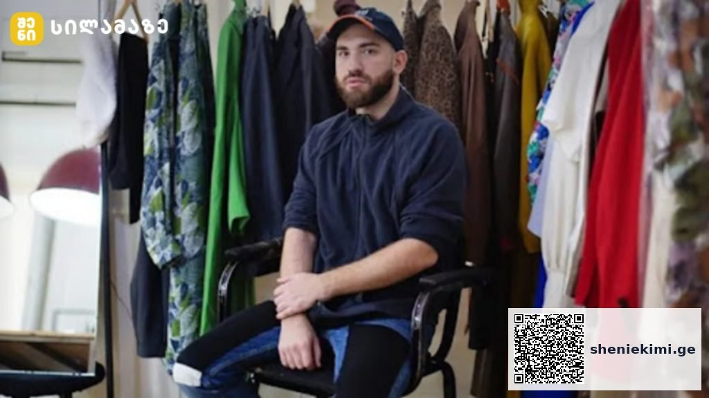 Bearded man wearing a navy hoodie and cap sits on a chair in a clothing studio surrounded by colorful garments on racks behind him.