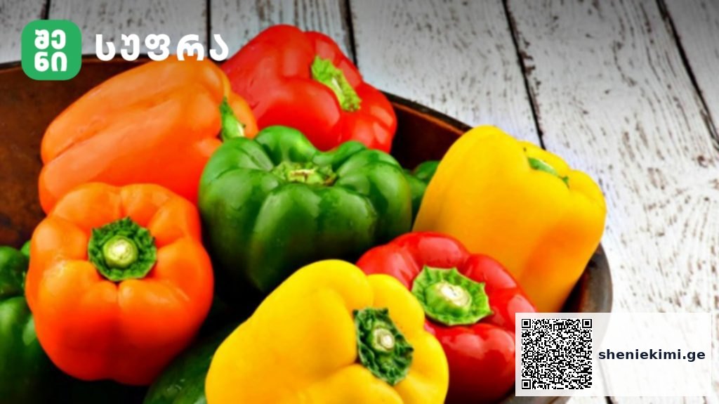 Assorted bell peppers (orange, green, red, yellow) in a bowl on a white wooden table, with a logo in the corner.