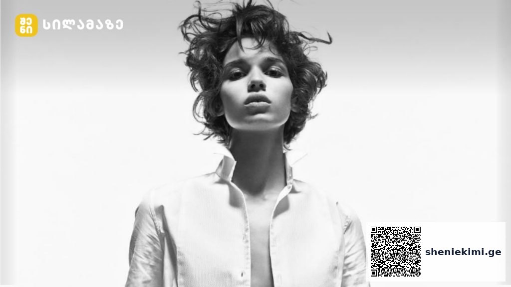 Black-and-white fashion portrait of an androgynous model in an open white shirt, looking straight ahead; logo top-left corner.