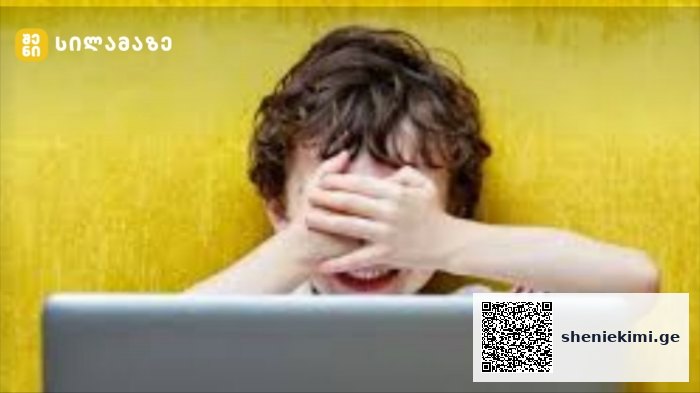Child covers eyes with hands while looking at a laptop in front of a yellow wall.