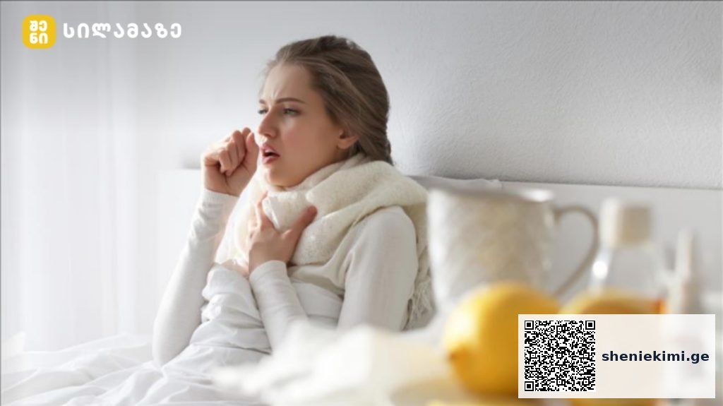 Woman in bed coughing into her elbow, wrapped in a scarf, with a blanket close by and bedside items like lemons and a teapot nearby.
