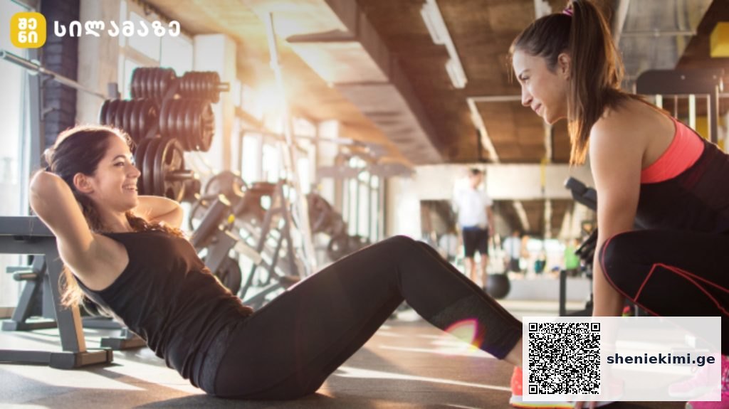 Two women workout in a gym: one lying on a mat doing sit-ups while the trainer crouches beside her offering support, with weights in the background.
