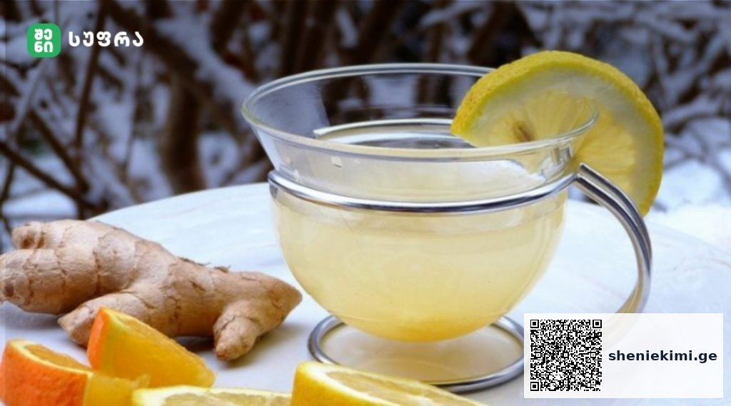 Glass mug of lemon-ginger tea with a lemon wedge on the rim, placed on a white surface with ginger root and orange pieces nearby.