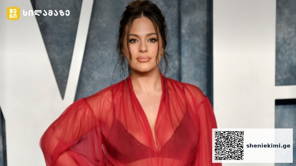 Woman wearing a sheer red dress posing on a red carpet at a formal event, dark hair pulled back and subtle makeup.