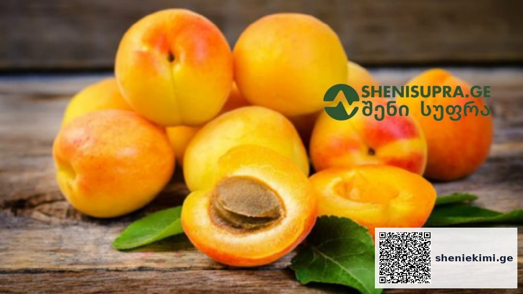 Pile of ripe apricots on a rustic wooden surface, with one cut open to show the stone and a couple of green leaves nearby in a natural setting.