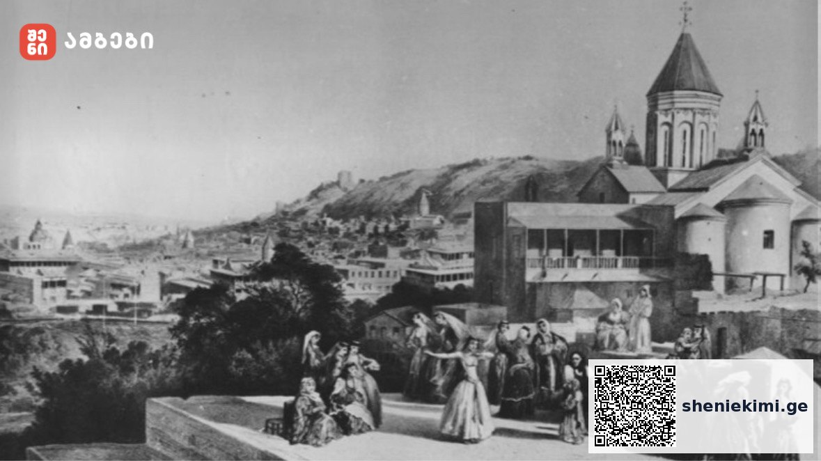 Black-and-white photo of a hillside town with a domed church and people in traditional clothing on a terrace.