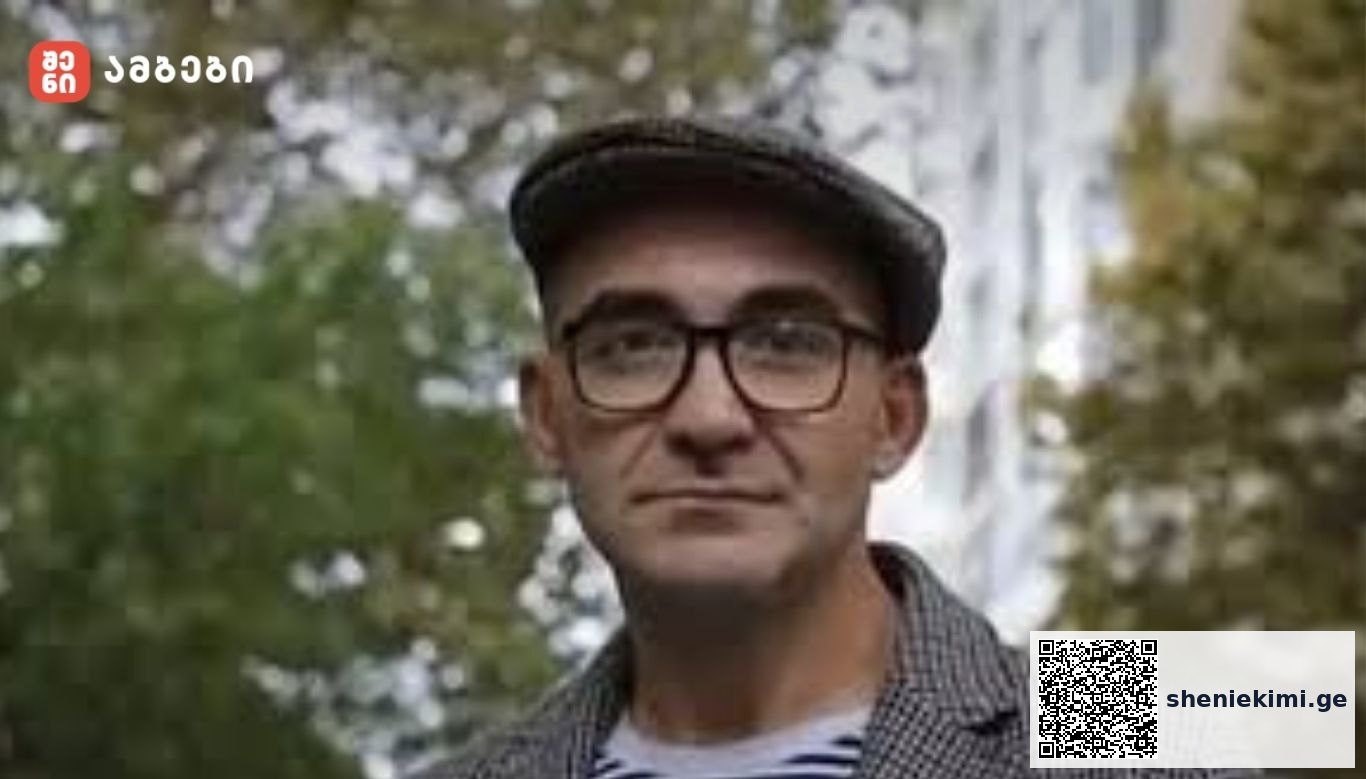 Portrait of a man wearing glasses and a flat cap outdoors, with a blurred fountain and greenery behind him; red logo in top-left corner.