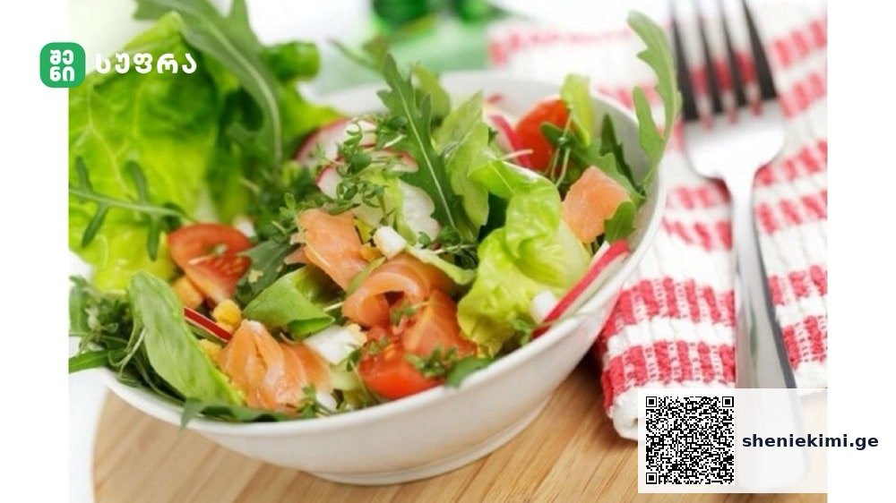 Bowl of mixed greens salad with tomatoes and smoked salmon, on a wooden table with a red-and-white striped napkin and fork nearby