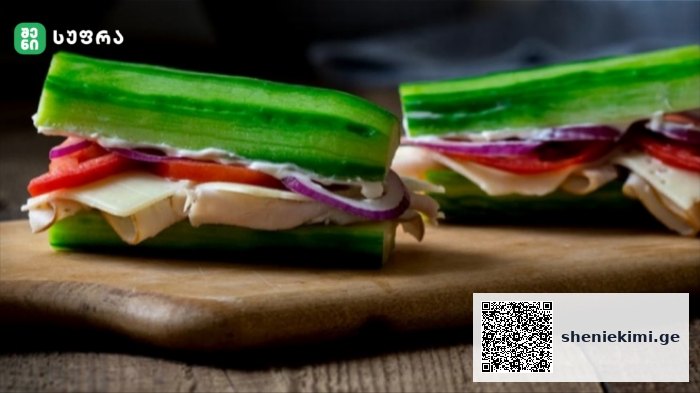 Two cucumber sandwiches filled with cheese, tomato, and red onion on a wooden cutting board.