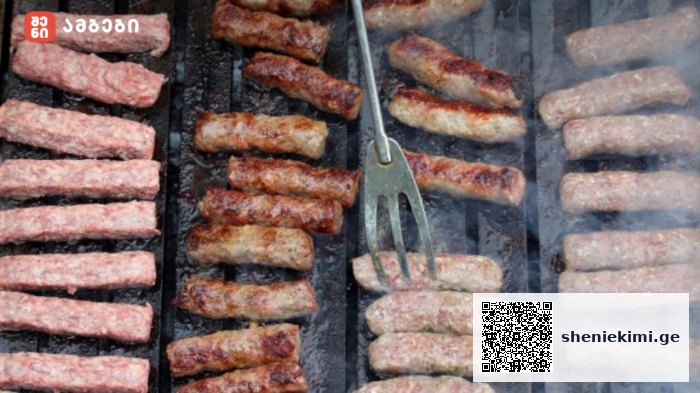 Close-up of sausage links grilling on a barbecue grate with smoky steam and a metal turning fork nearby.